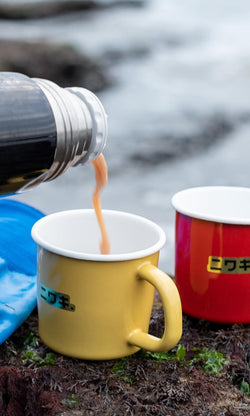 Plümo- A red enamel mug sits outdoors by water, next to a yellow cup as a hot drink is poured from a thermos.