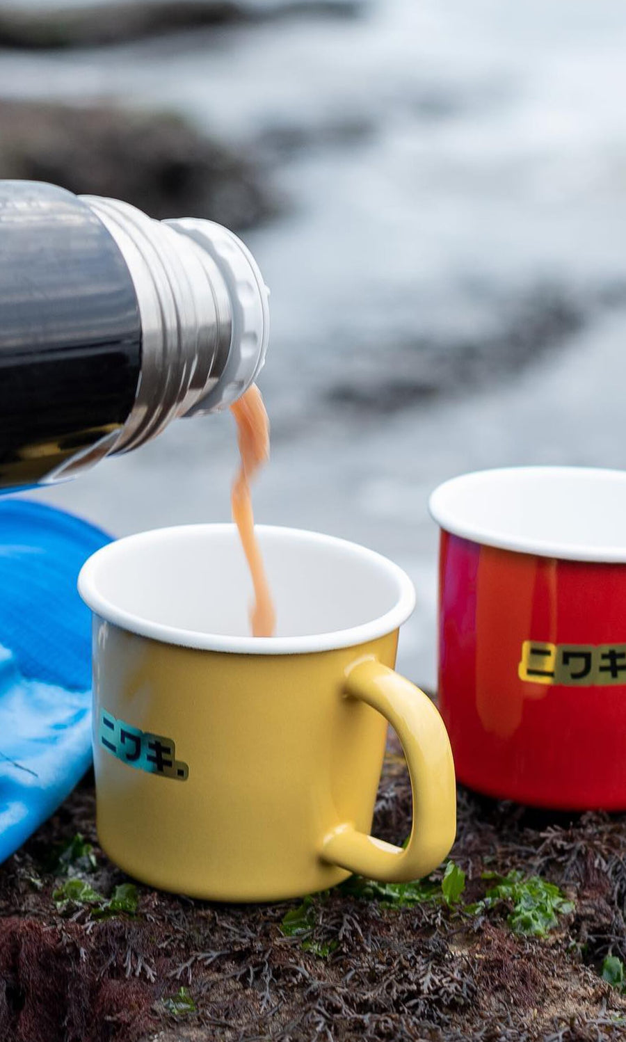 Plümo- A red enamel mug sits outdoors by water, next to a yellow cup as a hot drink is poured from a thermos.