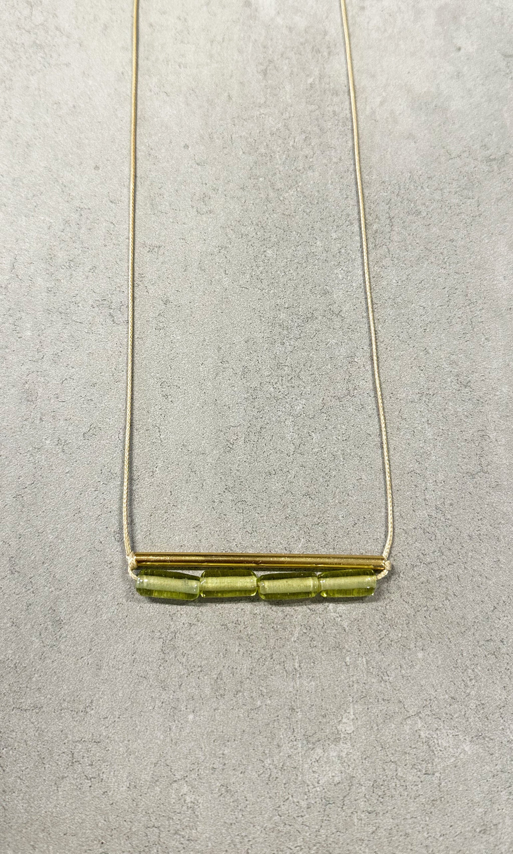 Plümo- The Lydia Necklace: a green beaded necklace with a gold bar and three glass beads on a light gray textured background.