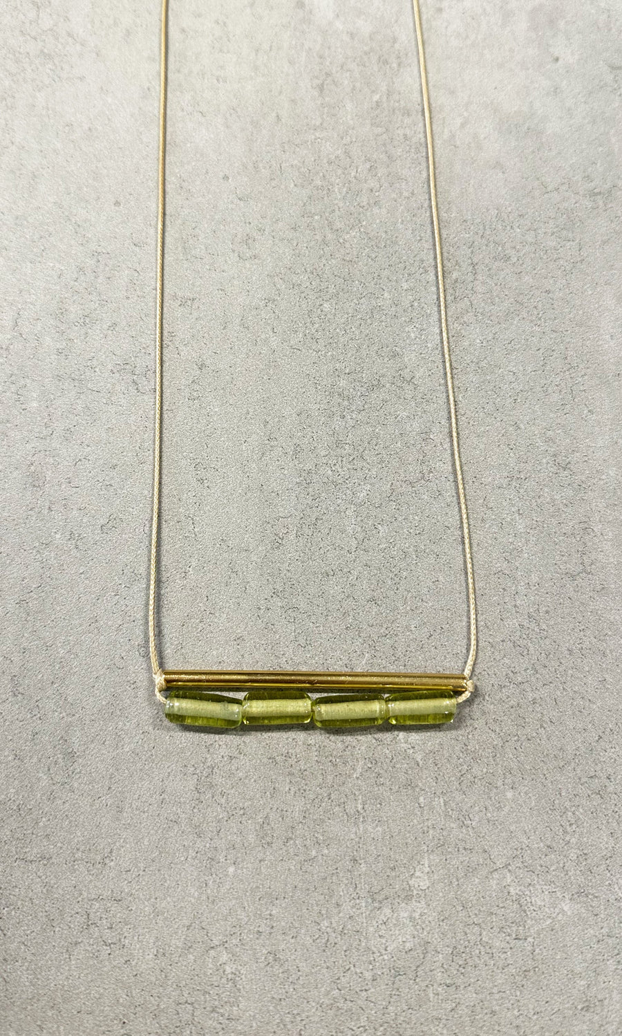 Plümo- The Lydia Necklace: a green beaded necklace with a gold bar and three glass beads on a light gray textured background.