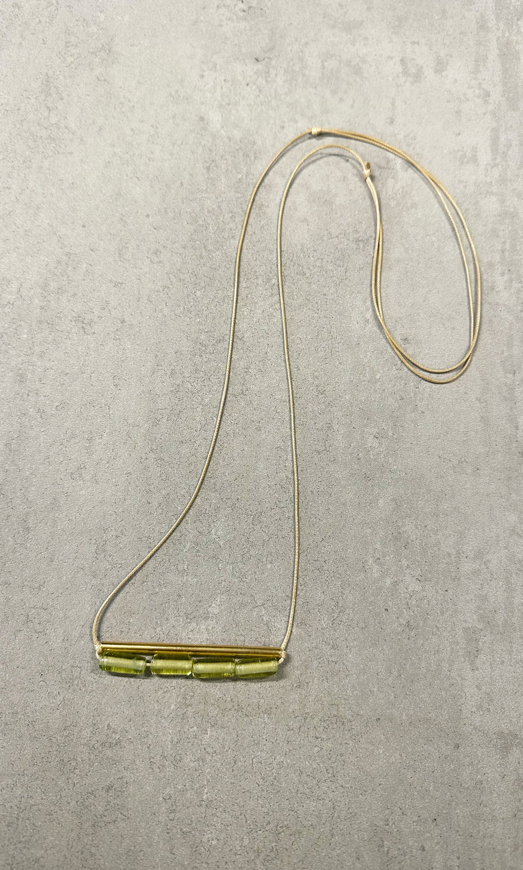 Plümo- The Lydia Necklace is a thin tan adjustable necklace with five green beads, shown on a light gray textured surface.