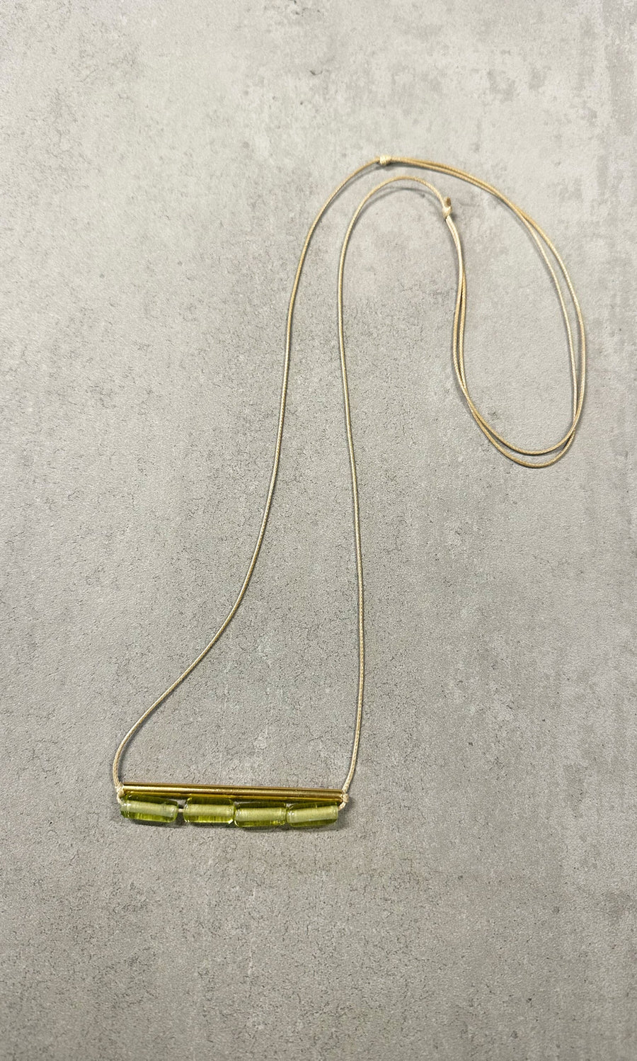 Plümo- The Lydia Necklace is a thin tan adjustable necklace with five green beads, shown on a light gray textured surface.
