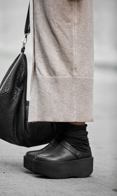 Plümo- A person wearing Kim Clogs, black socks, a beige long skirt, and holding a large black textured bag.