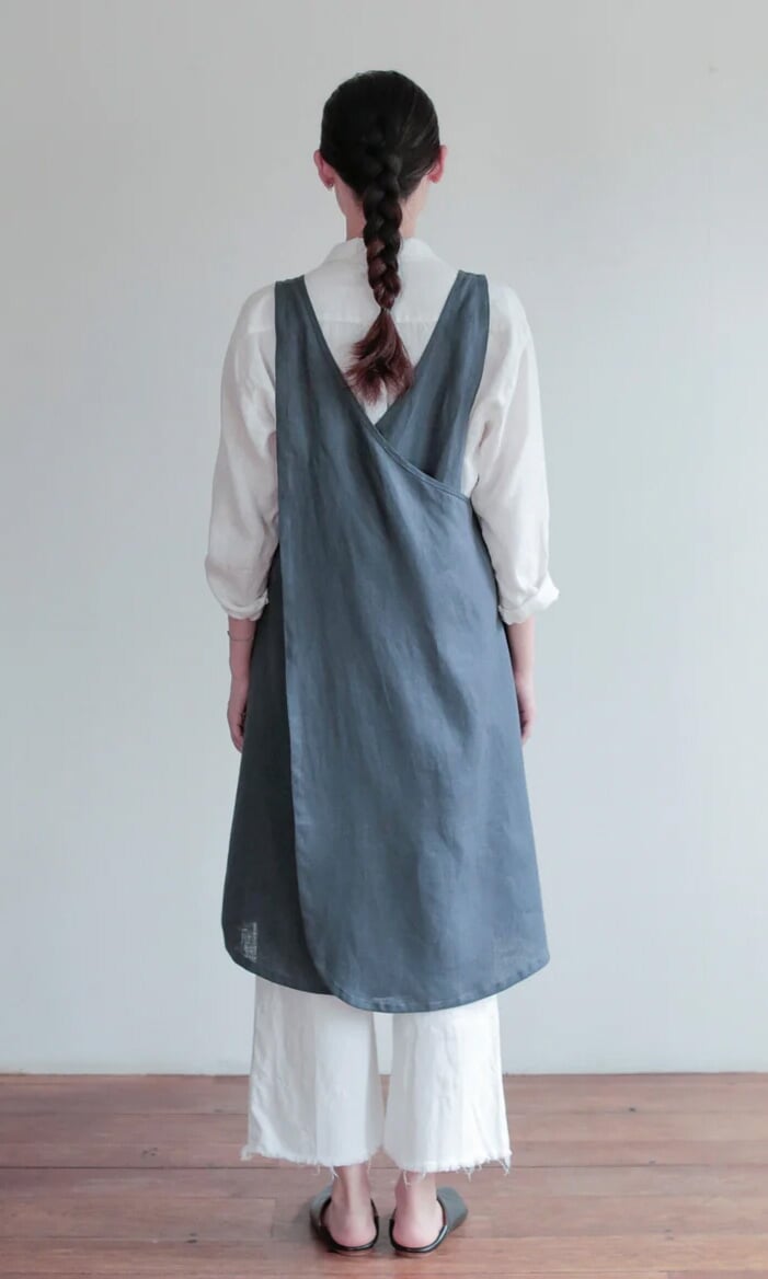 Plümo- A person faces away, wearing the Arden Pinafore blue linen cross-back apron over a white shirt and wide white pants.