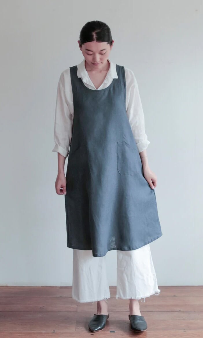 Plümo- A woman in the Arden Pinafore, a blue linen cross-back apron, stands on a wooden floor in a white shirt and pants.
