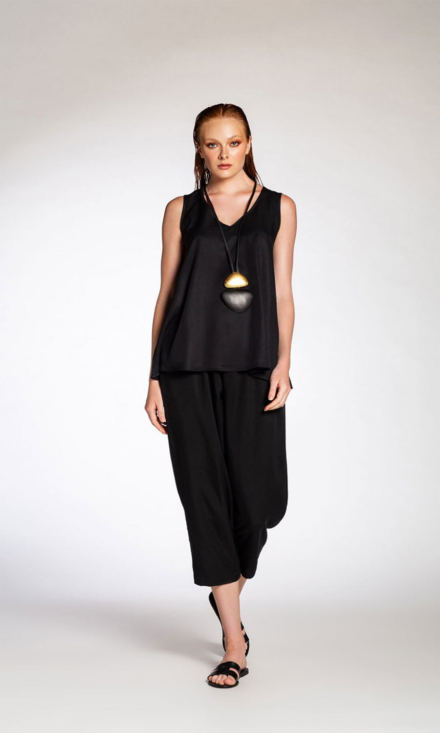 Plümo- Woman wears the Carmen Top - Tencel with loose black pants, sandals, and a pendant necklace against a plain background.
