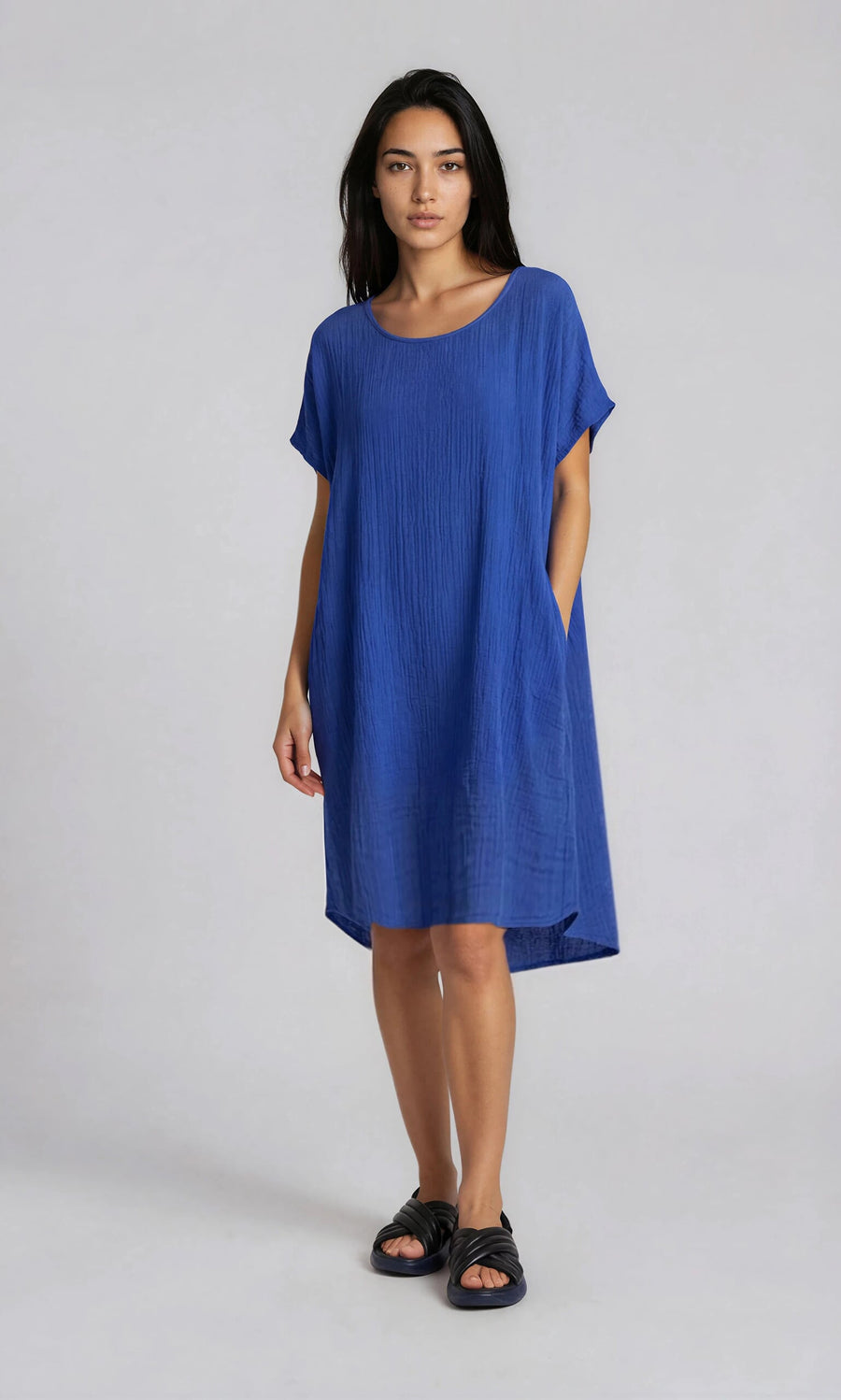 Plümo- Woman in the Eleni Dress, a loose, sheer blue beach dress with oversized pockets and black sandals, on plain backdrop.