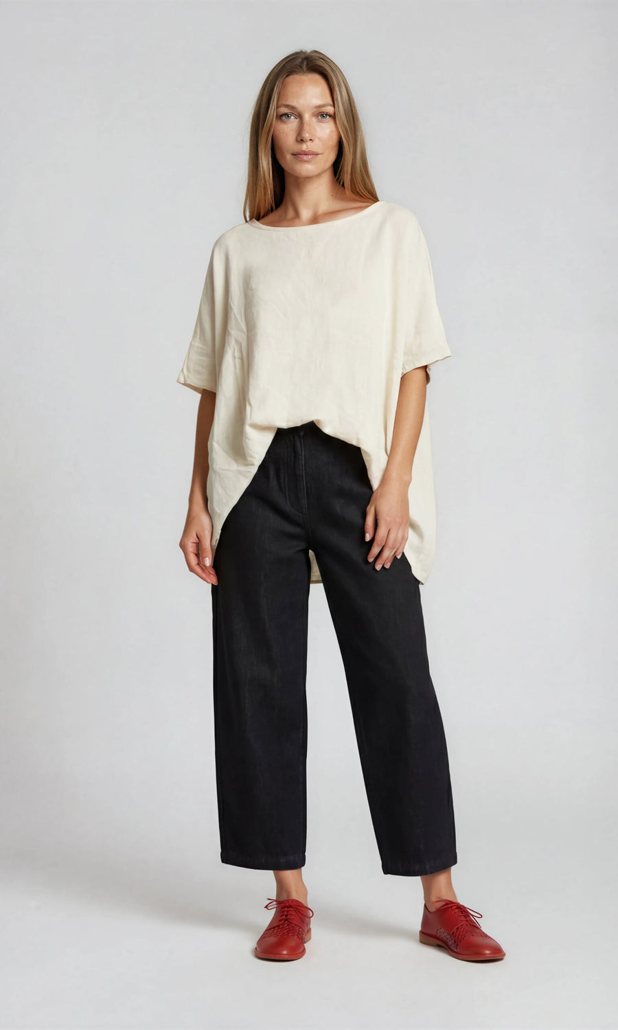 Plümo- Woman wears the oversized, boxy Marielle Top in ecru, black wide-leg pants, and red shoes against a plain background.