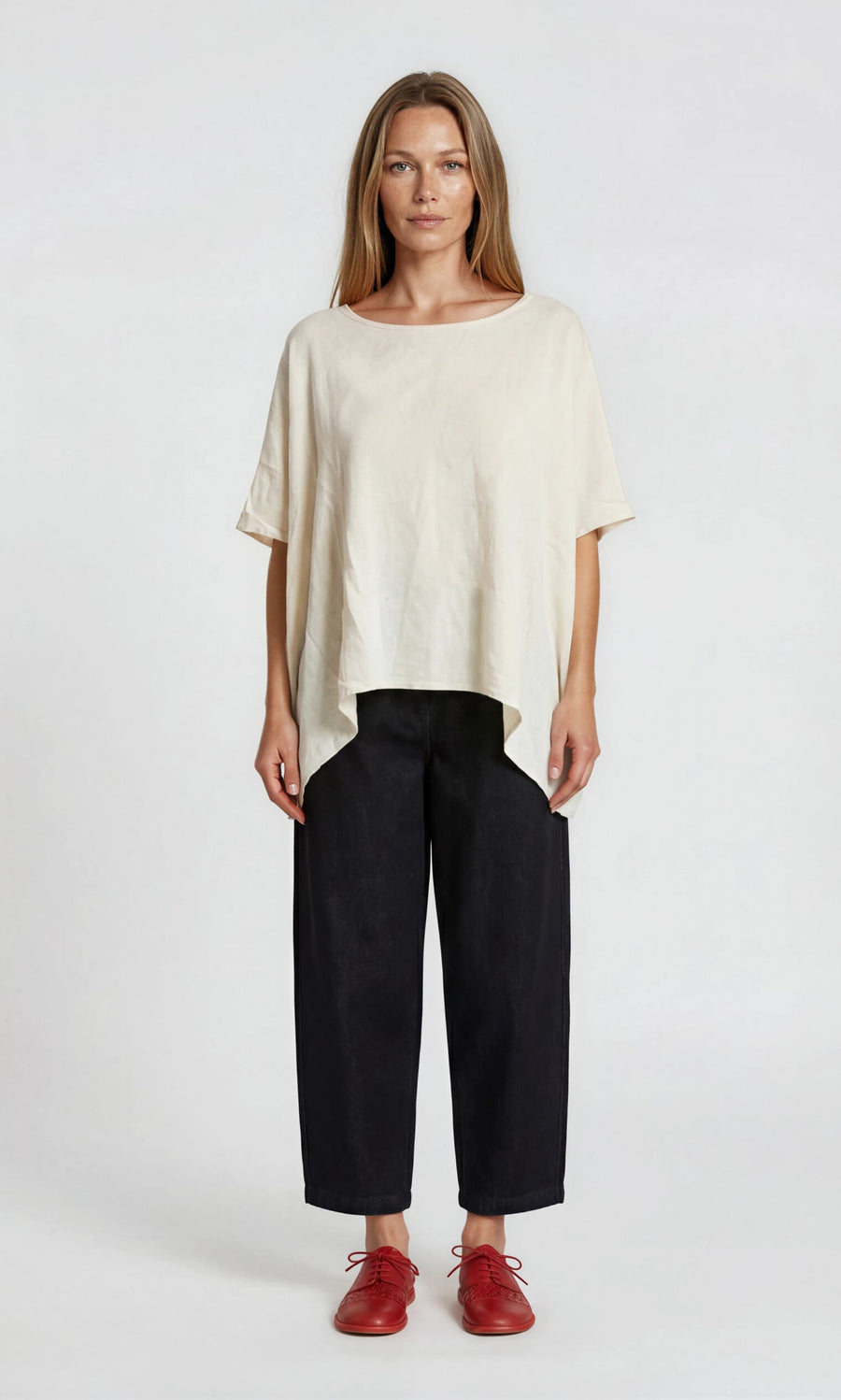 Plümo- Woman in an oversized Marielle Top ecru, black pants, and red shoes against a white background.