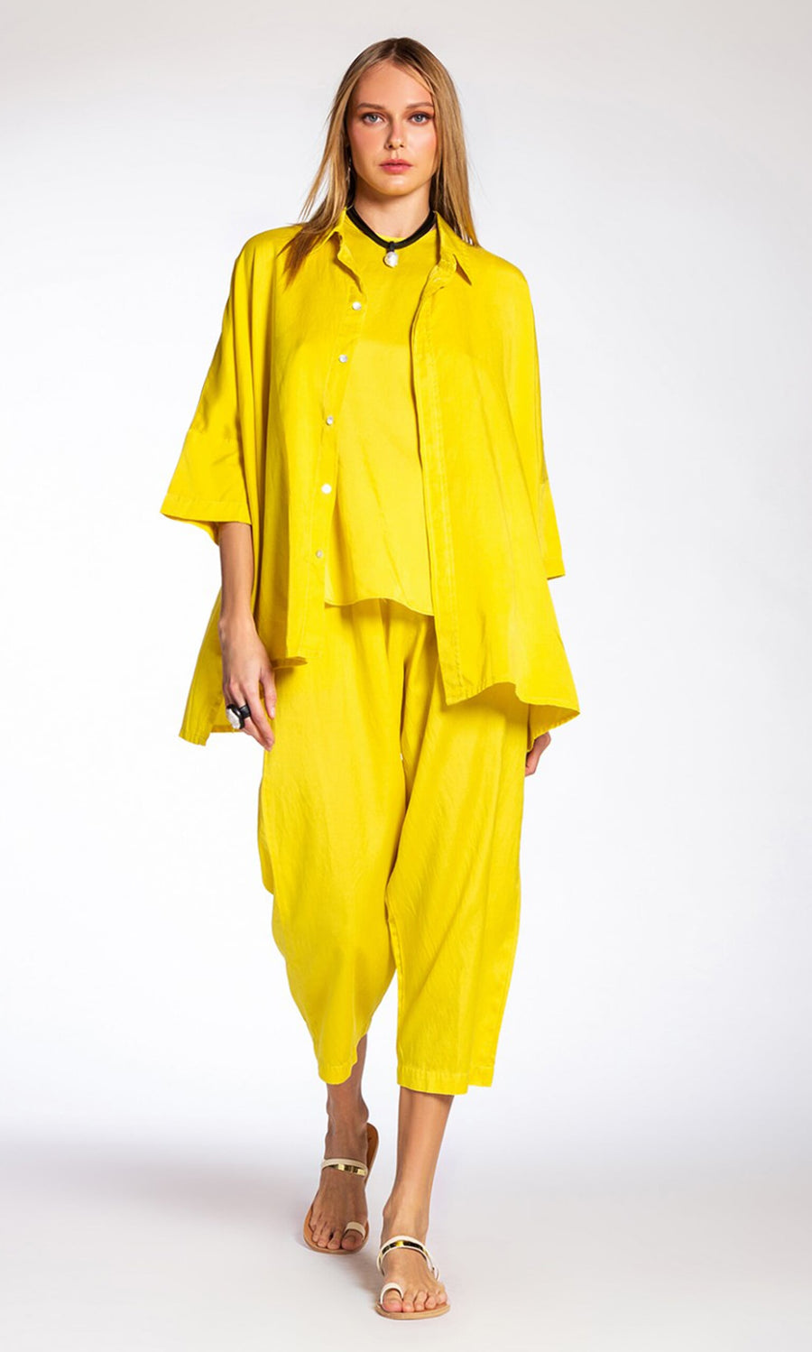 Plümo- Woman models Marigold Pants—relaxed fit, yellow and silky—with a bright shirt and white sandals against a plain backdrop.