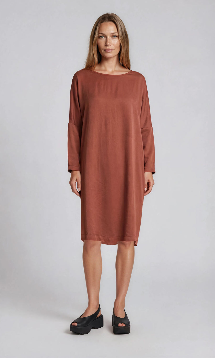 Plümo- A woman wears the Remi Dress in rust silk with black platform sandals, posing against a plain background.