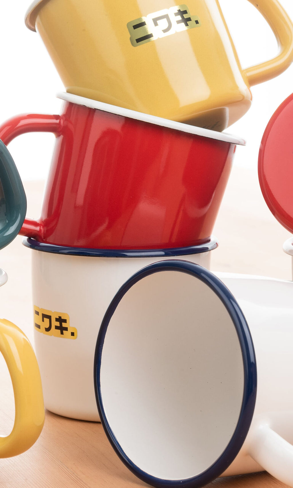 Plümo- Red enamel mug displayed on a wooden surface with stacked yellow, white, and Japanese-script mugs nearby.