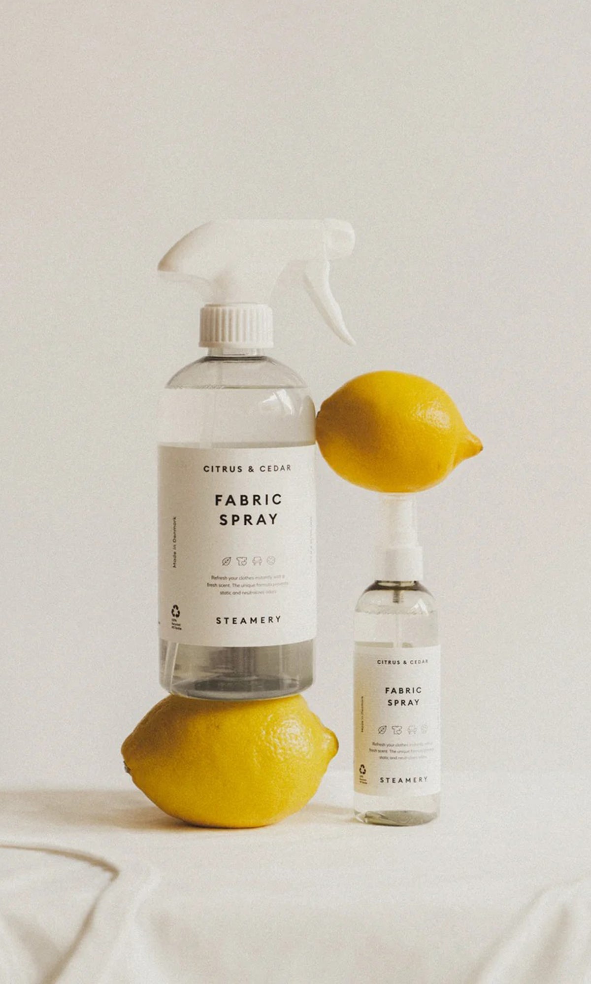 Plümo  Two lemons and two Citrus  Cedar Fabric Spray 500ml bottles rest on a sleek white surface and background.