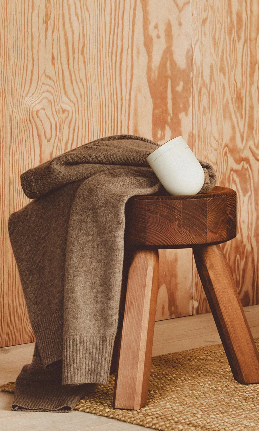 Plümo  A folded brown sweater for the Fabric Groomer, plus a white mug, sit on a wooden stool by a woven rug.