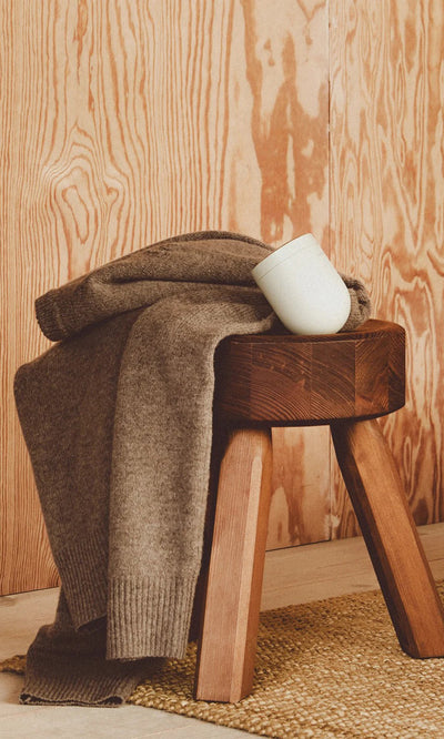 Plümo  A folded brown sweater for the Fabric Groomer, plus a white mug, sit on a wooden stool by a woven rug.