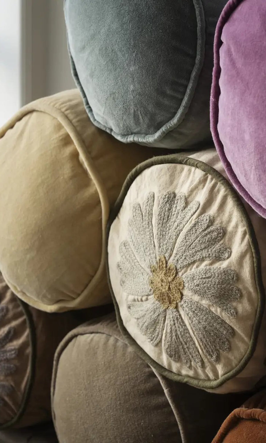 Plümo- Stack of round cushions in soft colors, featuring the Velvet Bolster Cushion with detailed floral embroidery on the front.