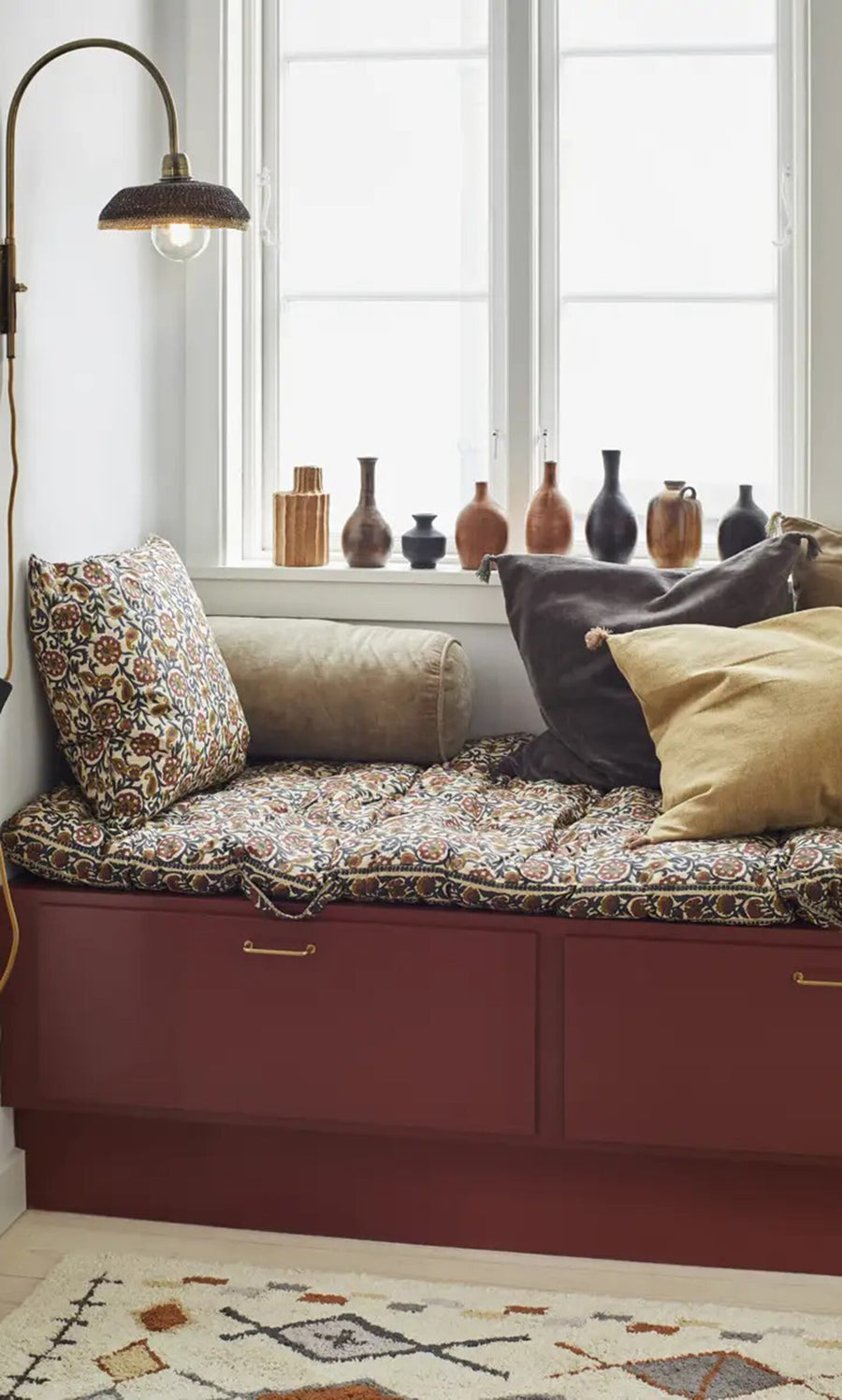 Plümo- A cozy window seat with a Velvet Bolster Cushion and vases on the sill beneath a brass wall lamp.